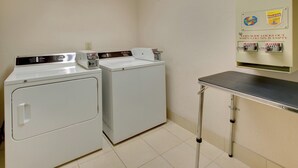 A laundry area with two front-loading washers, a coin-operated dryer, and a metal utility table. - Holiday Inn Express Hotel & Suites Pleasant Prairie-Kenosha by IHG (Pleasant Prairie)