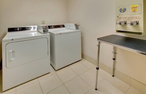 A laundry area with two front-loading washers, a coin-operated dryer, and a metal utility table. - Holiday Inn Express Hotel & Suites Pleasant Prairie-Kenosha by IHG (Pleasant Prairie)