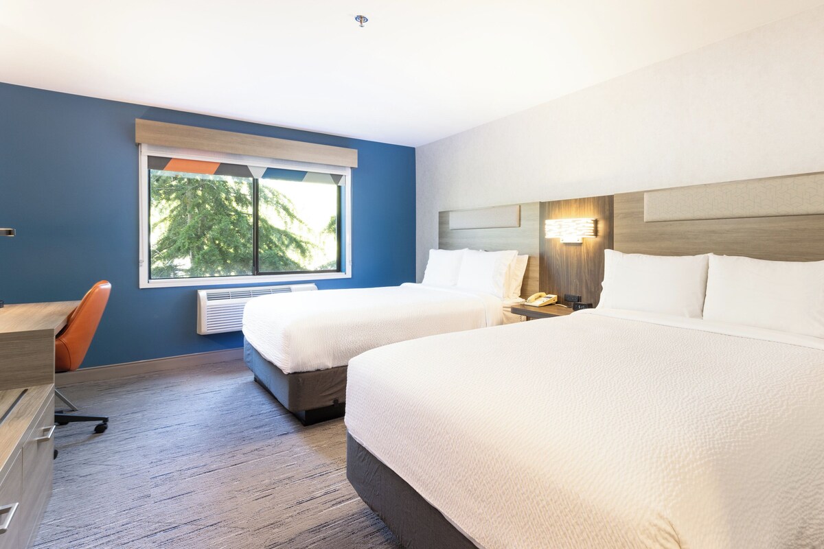 A hotel room with two beds, a desk, and a window with a view of trees.