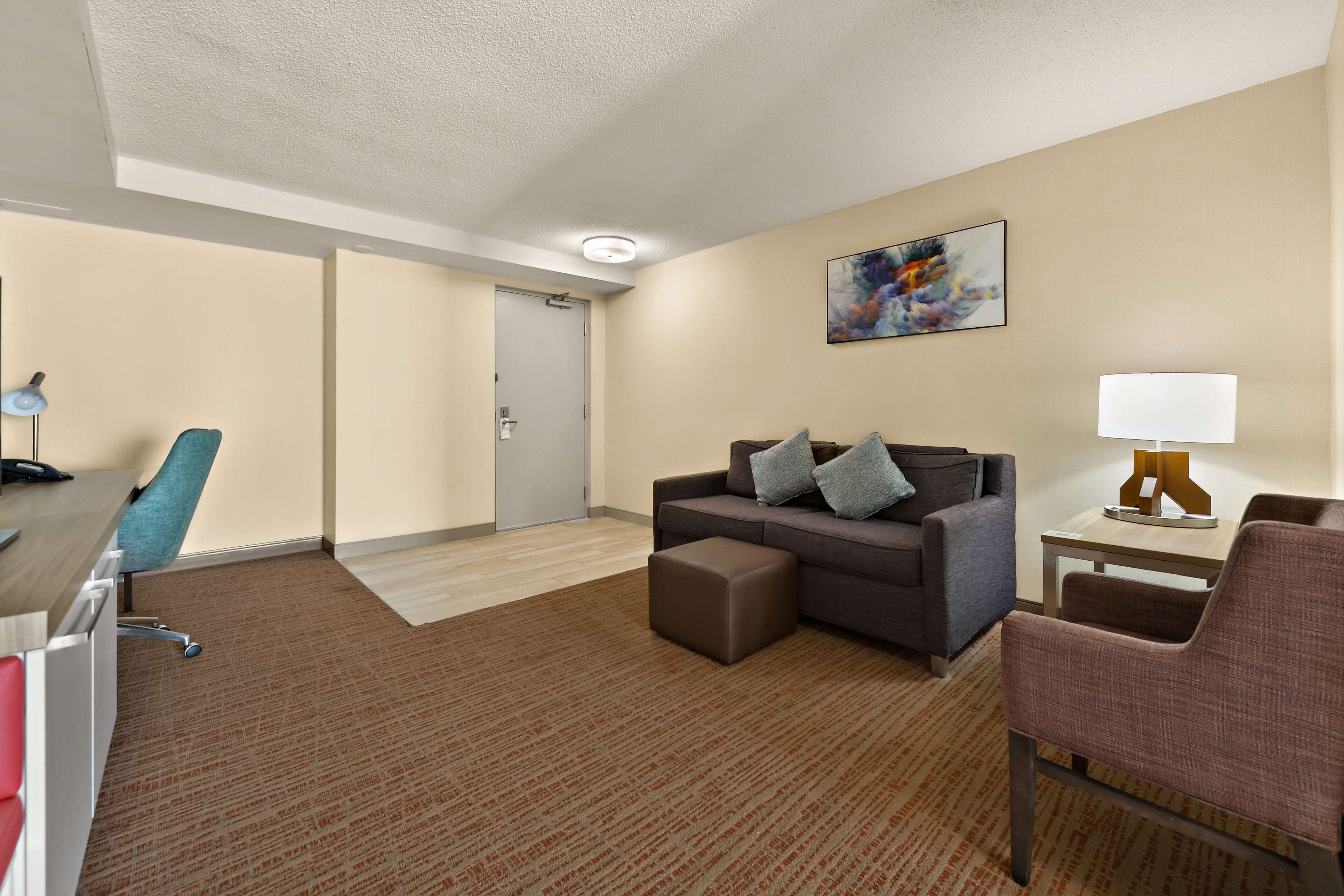 A hotel room with a brown carpet, a gray sofa with pillows, a blue chair, a wooden desk with a lamp, and a painting on the wall.