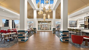 A spacious lobby with high ceilings, modern pendant lighting, and a central seating area with red chairs. - Hilton Garden Inn Williamsburg (Williamsburg)