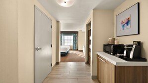 A hotel hallway with a wooden floor, a white door, a kitchen area with a microwave and coffee maker, and a bed with blue curtains in the background. - Hilton Garden Inn Williamsburg (Williamsburg)