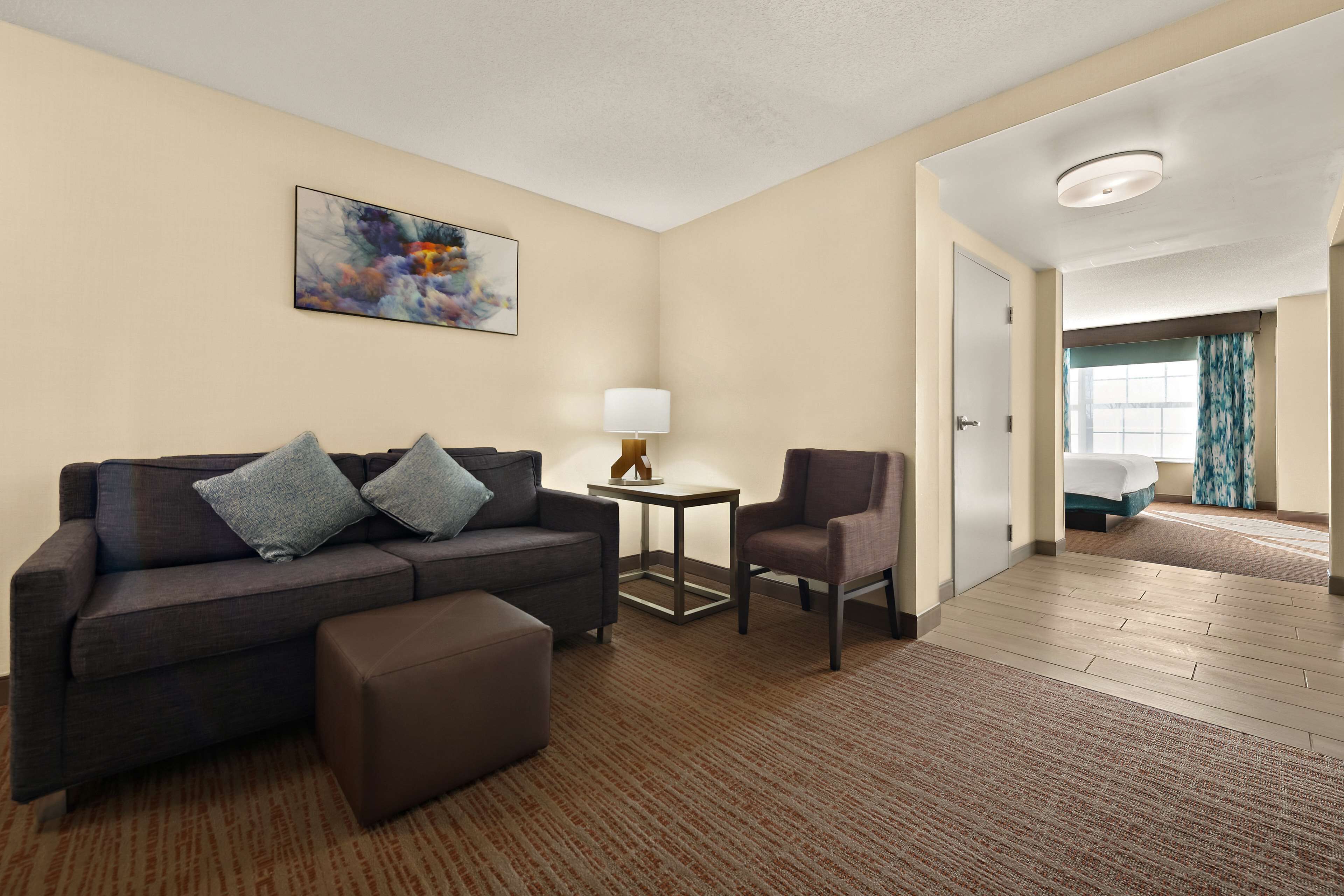 A hotel room with a dark brown sofa, a small side table with a lamp, a chair, and a large window with curtains.