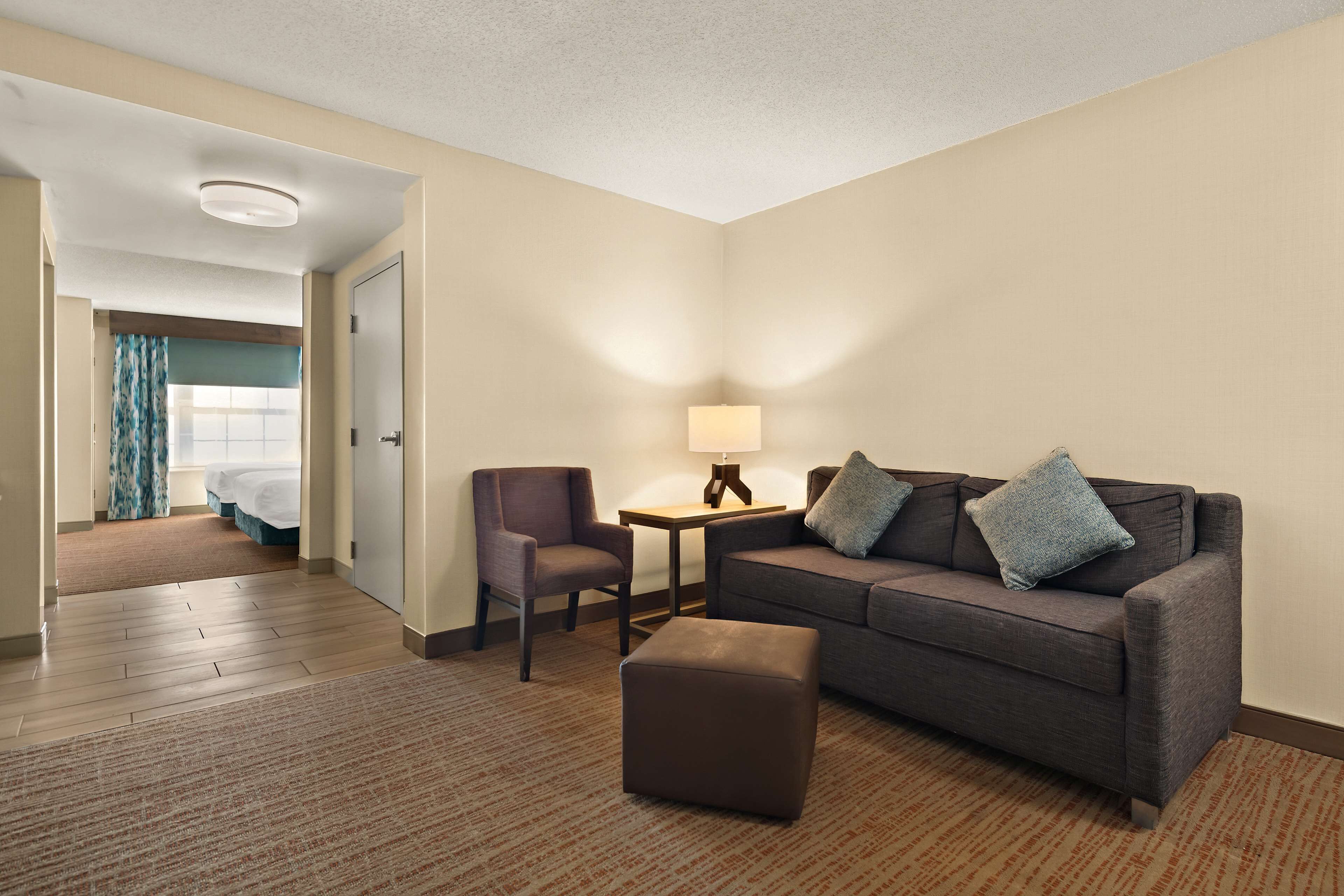 A hotel room with a sofa, armchair, ottoman, and a small table with a lamp.