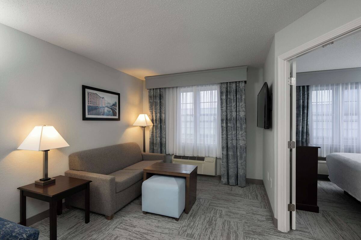 A hotel room with a sofa, a small table, a lamp, a picture on the wall, a bed, and a window with curtains.