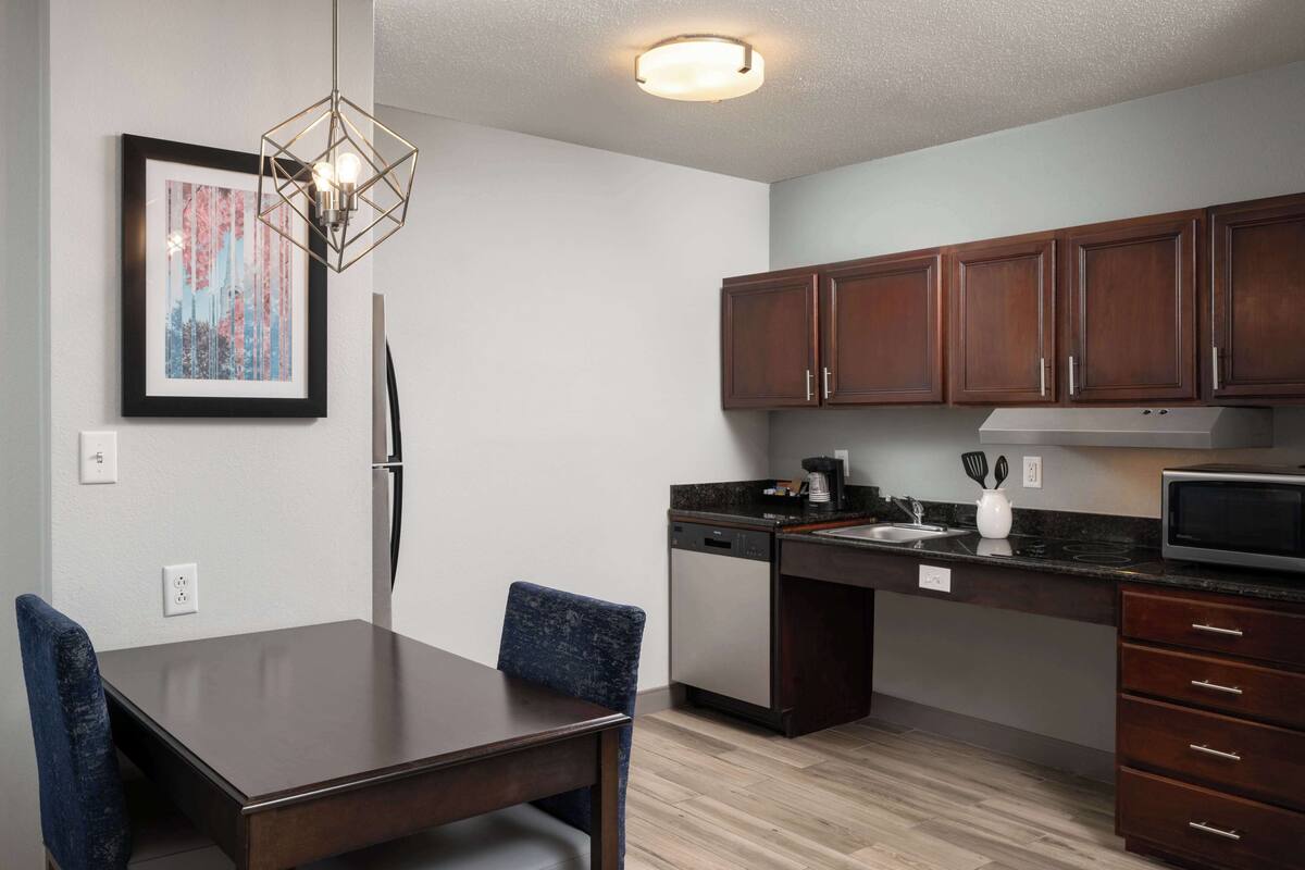 A modern kitchen with dark wooden cabinets, a built-in dishwasher, and a microwave. There is a dining table with chairs and a pendant light above it.
