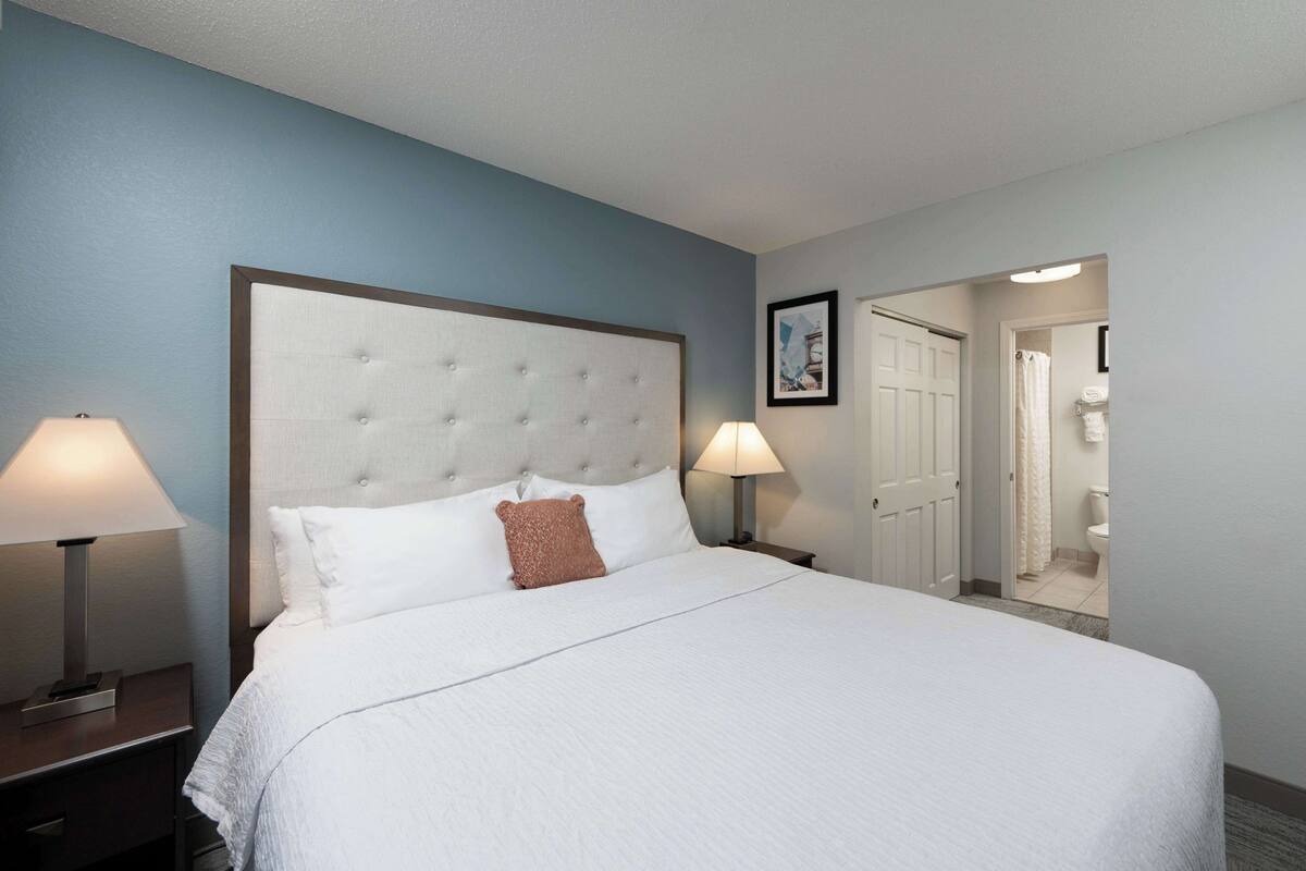 A hotel room with a large bed, two bedside lamps, a framed picture on the wall, and a door leading to a bathroom.
