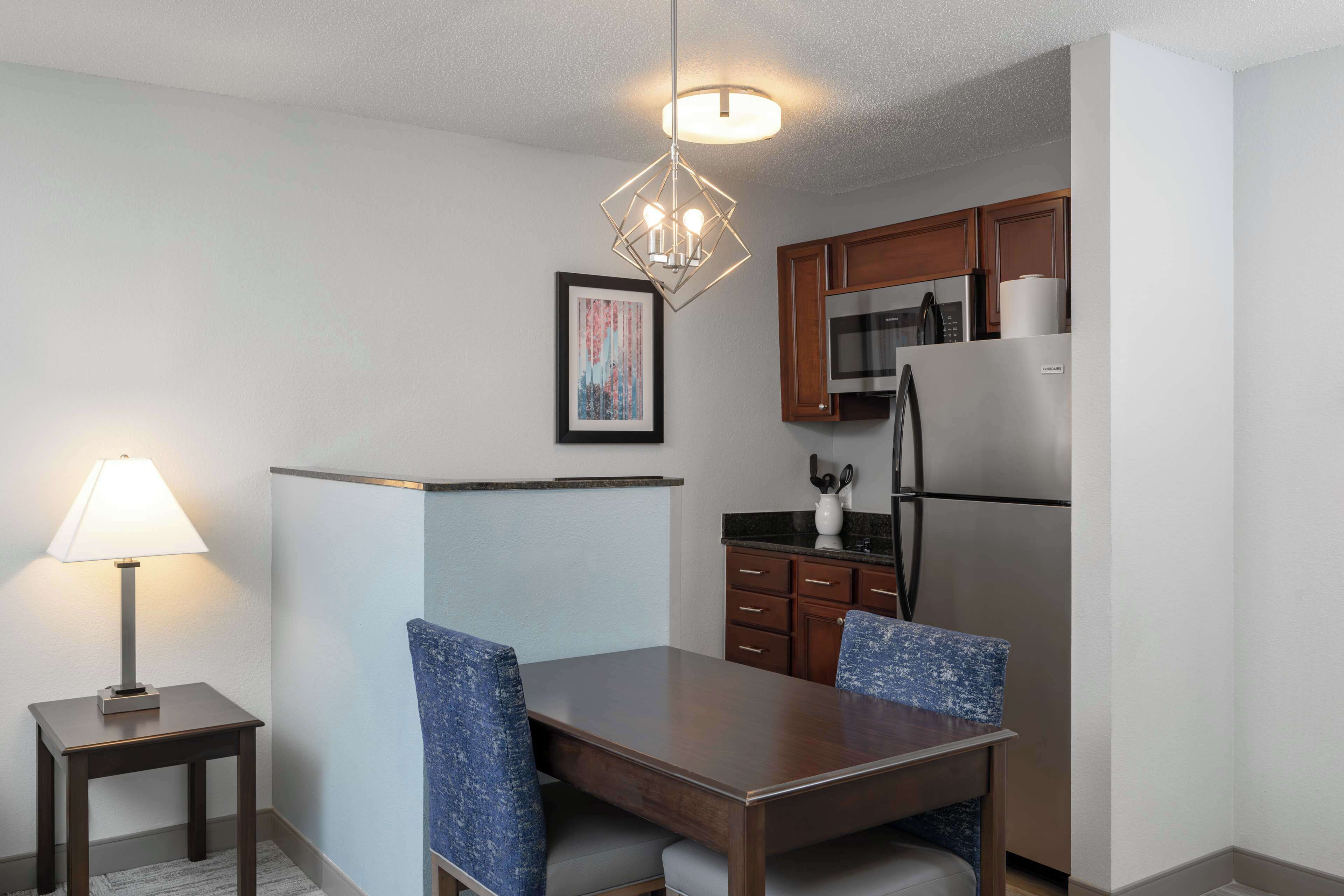 A modern kitchen with a refrigerator, microwave, and wooden cabinets. There is a dining table with two chairs and a lamp on a side table.
