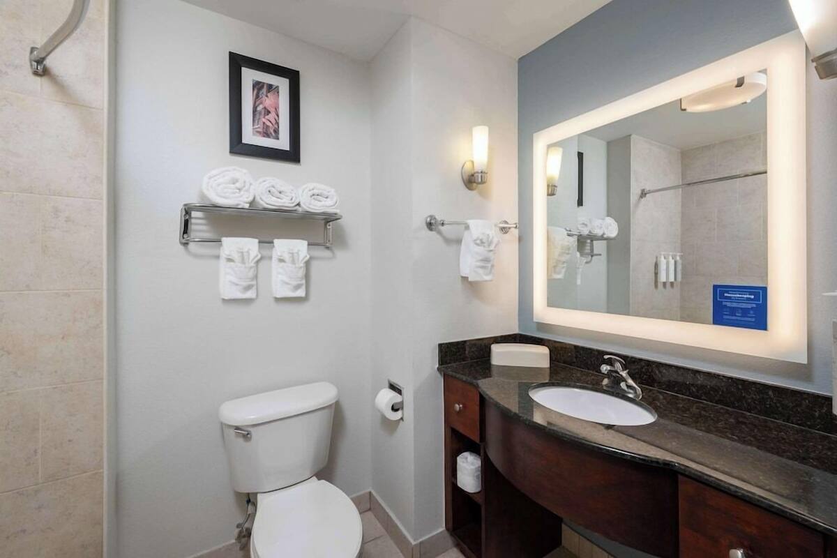 A modern bathroom with a toilet, sink, and a large mirror. There are towels on a shelf and a framed picture on the wall.