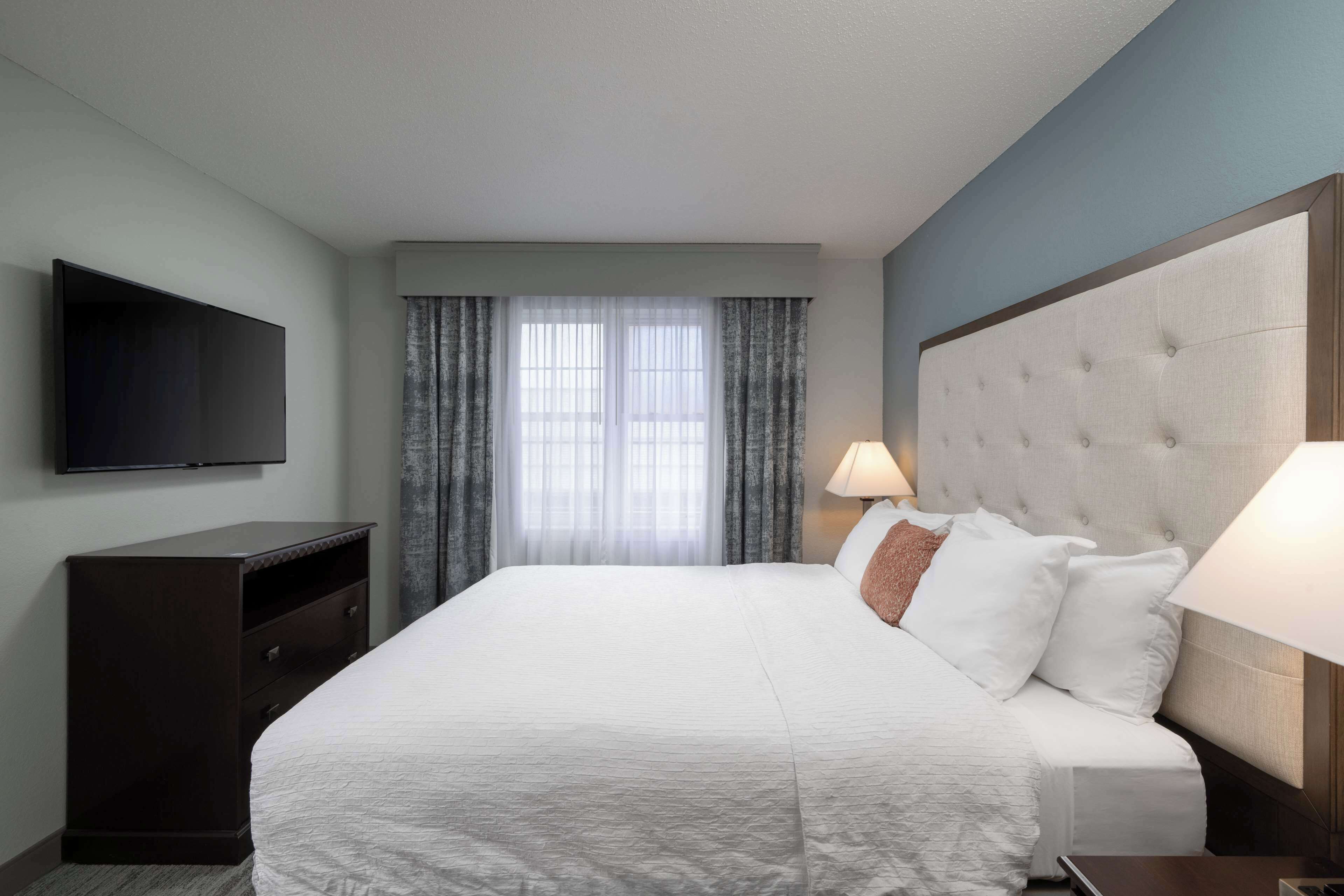 Habitación de hotel con una cama grande, un televisor de pantalla plana en la pared, una mesita de noche con lámpara y una ventana con cortinas.