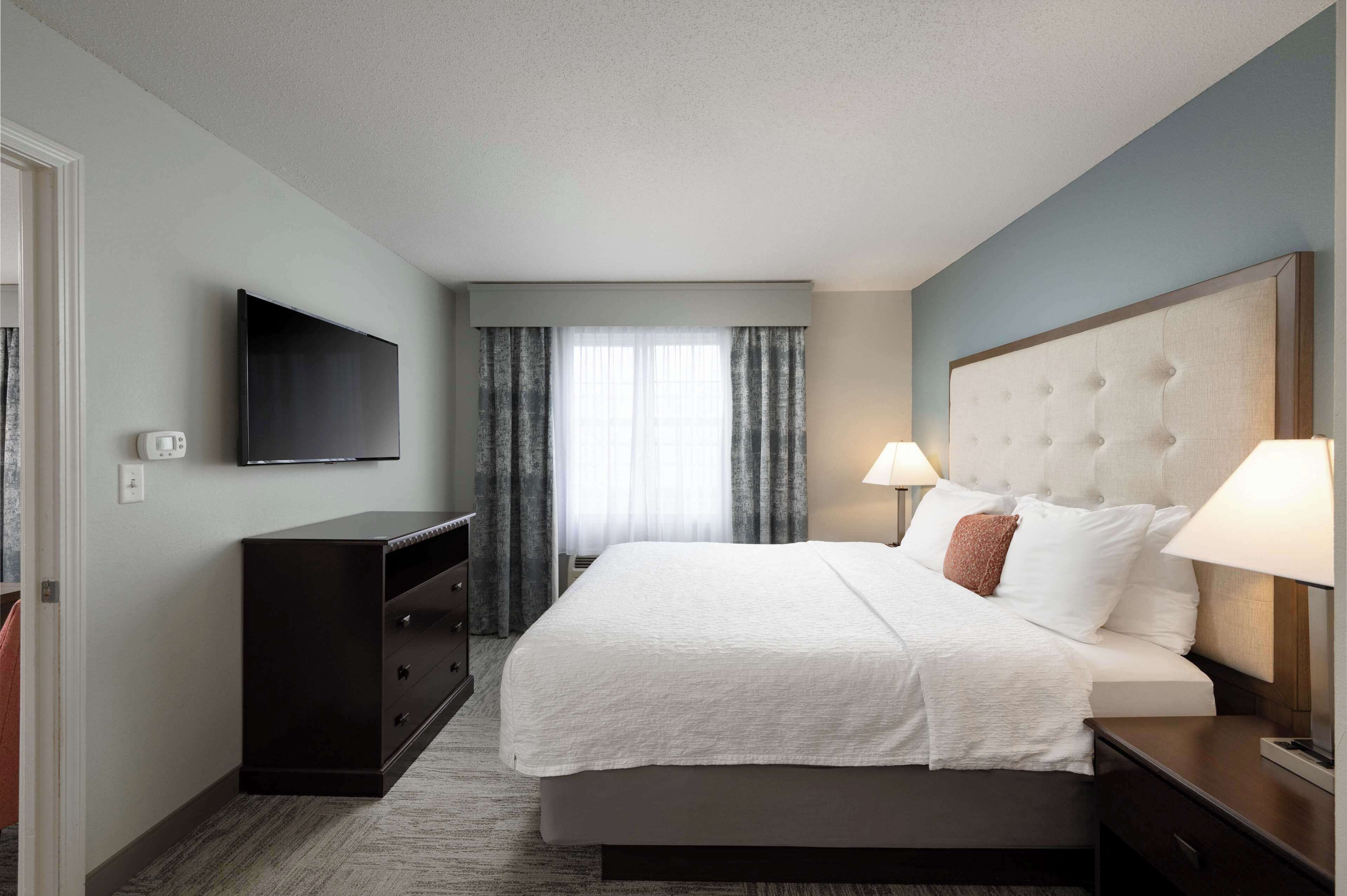 A hotel room with a large bed, a flat-screen TV mounted on the wall, a dresser, and a window with curtains.