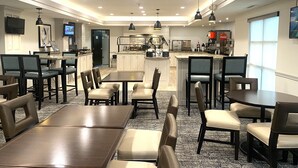 Un espace repas moderne avec plusieurs tables et chaises, une cuisine à l’arrière-plan et un téléviseur fixé au mur.