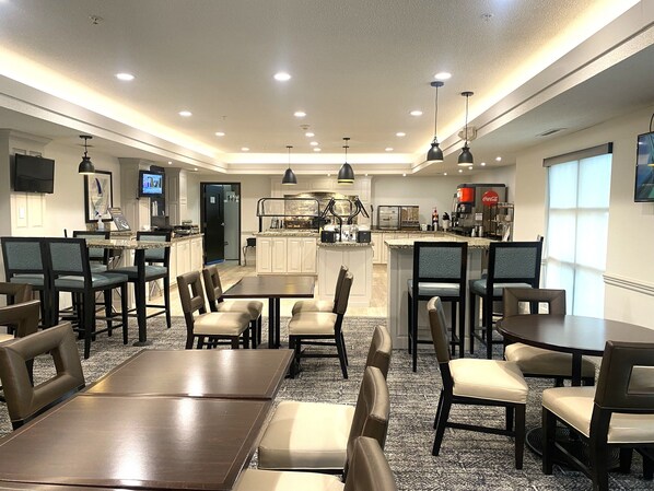 A modern dining area with multiple tables and chairs, a kitchen in the background, and a television mounted on the wall. - Staybridge Suites Columbia by IHG (Columbia)