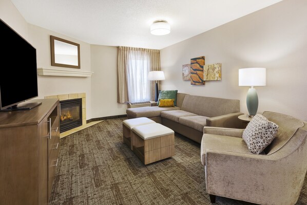 A hotel room with a flat-screen TV, a fireplace, a sofa, a loveseat, and a side table with a lamp.