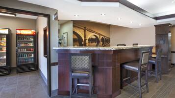 A bar area with a counter and two bar stools, a refrigerator with beverages, and a wall-mounted picture of a bridge.