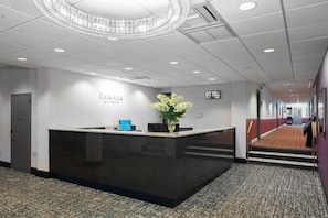 Un mostrador de recepción con un reloj marcando las 07:10, un arreglo floral y un letrero que dice 'RAMADA BY WYNDHAM'.