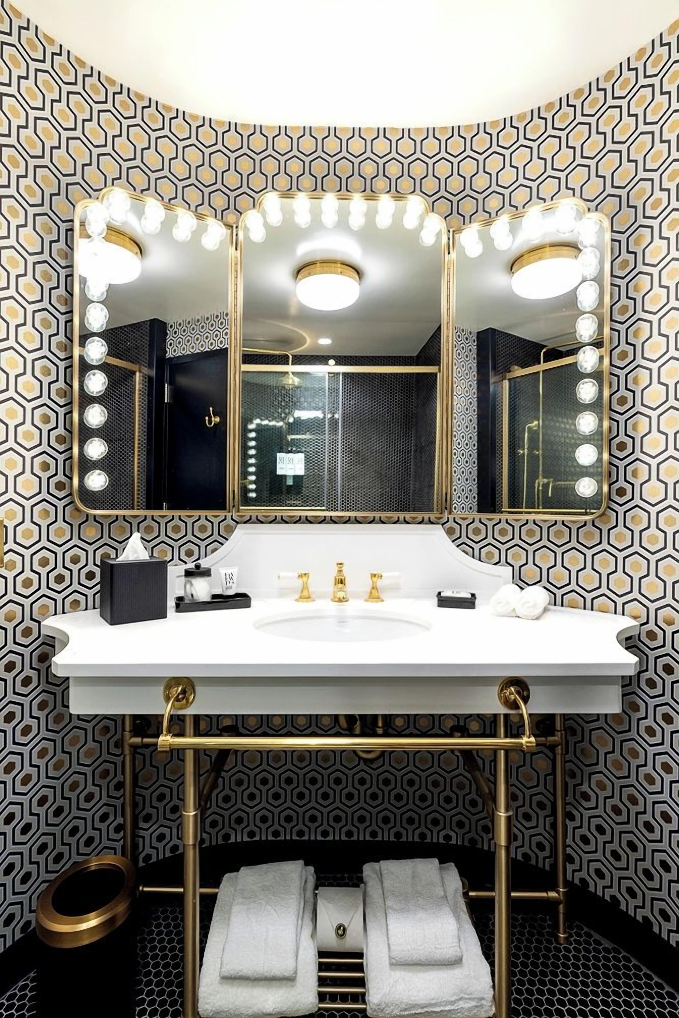 A bathroom with a vanity, three mirrors with lights, a gold faucet, and a patterned wall.