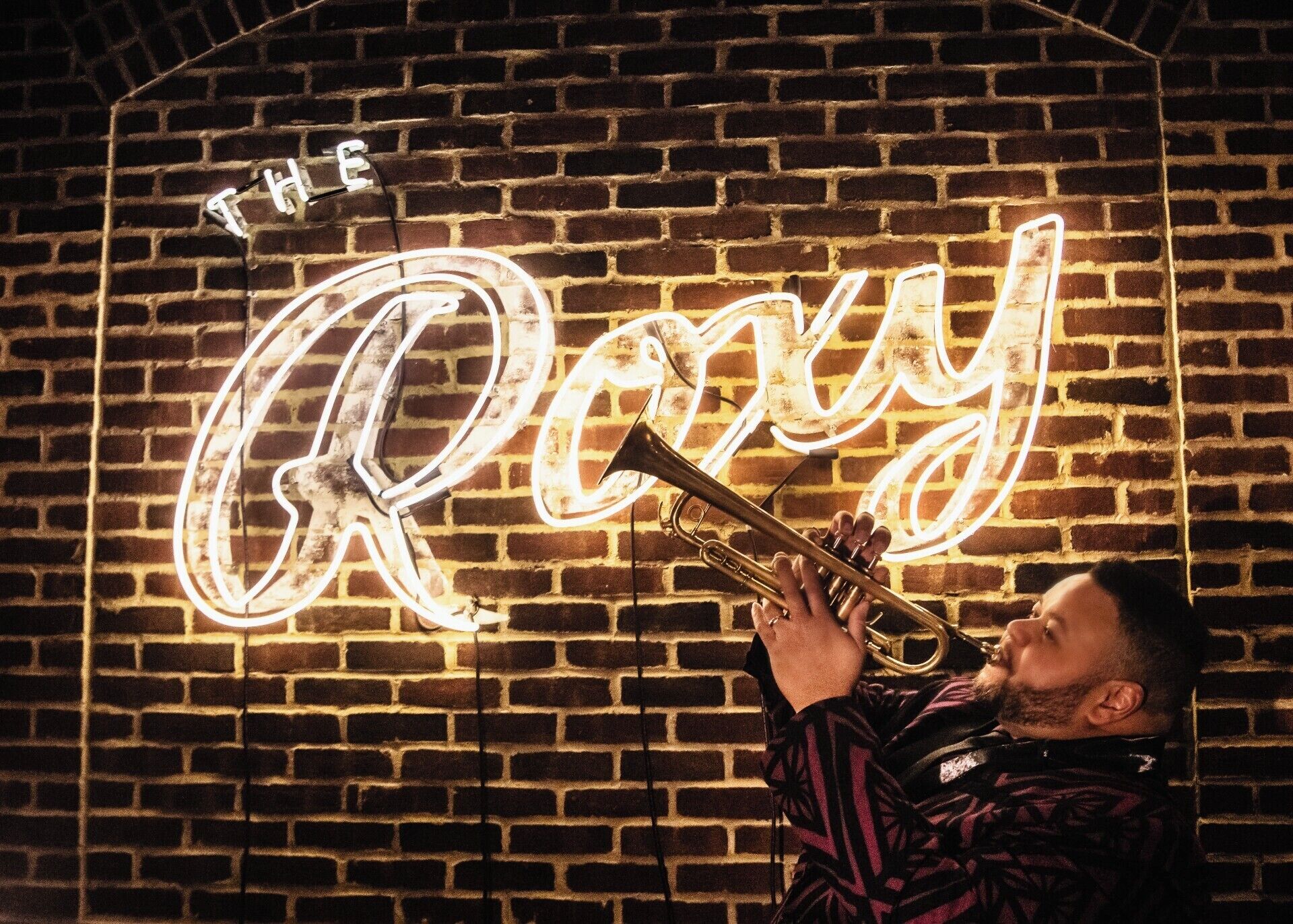 Eine Person spielt vor einem Neon-Schild, auf dem 'THE Roy' steht, Trompete.