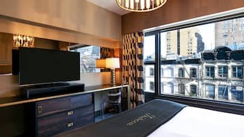 Quarto de hotel moderno com uma cama grande, televisão de tela plana, escrivaninha com cadeira e vista para a cidade através de amplas janelas.