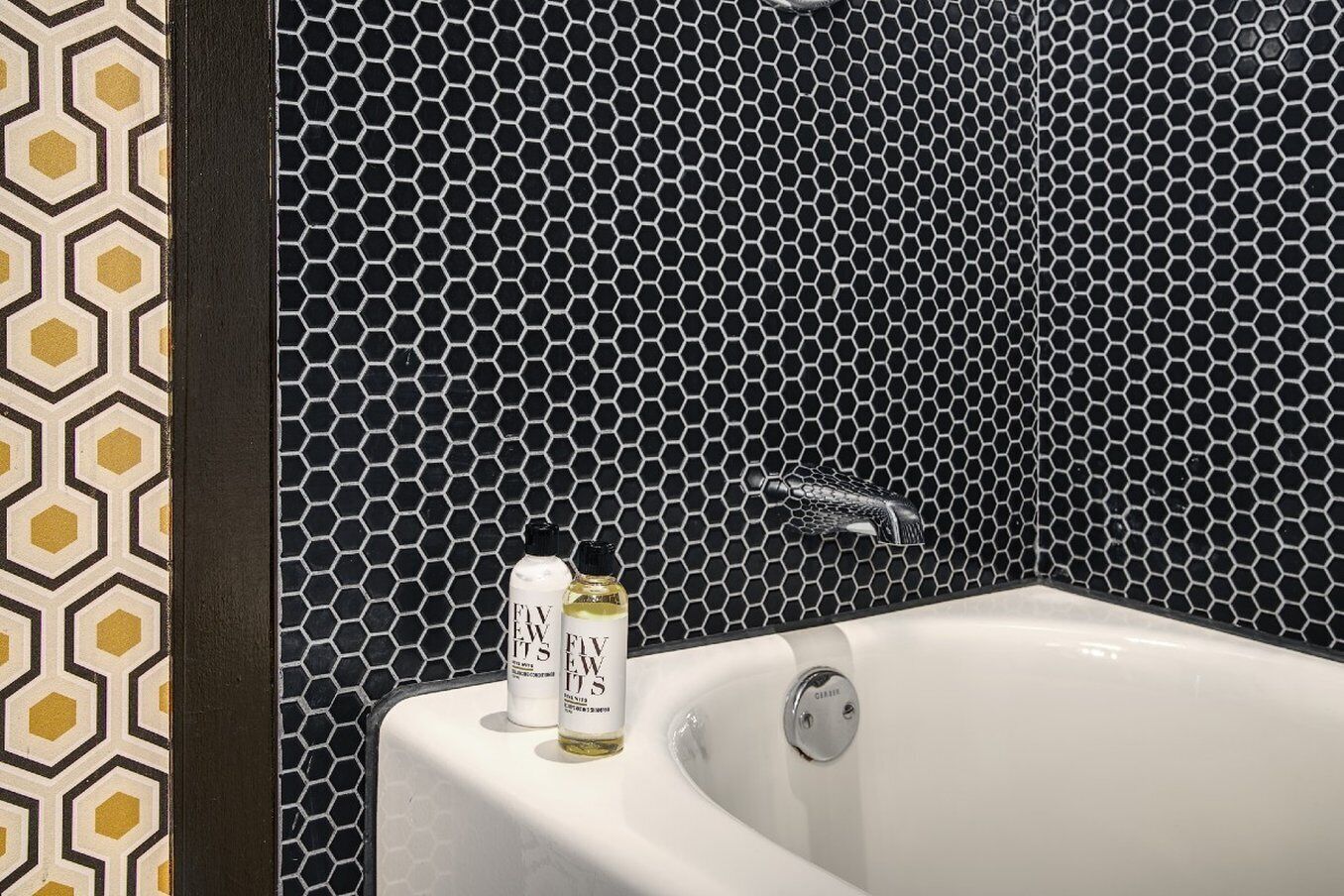 Een badkamer met een betegeling in een honingraatpatroon, een witte badkuip en twee flessen 'Five Weeks' shampoo.