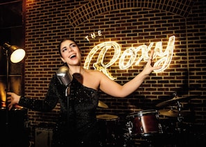 A person in a black outfit with a microphone, standing in front of a brick wall with a neon sign that reads 'THE Roxy'.