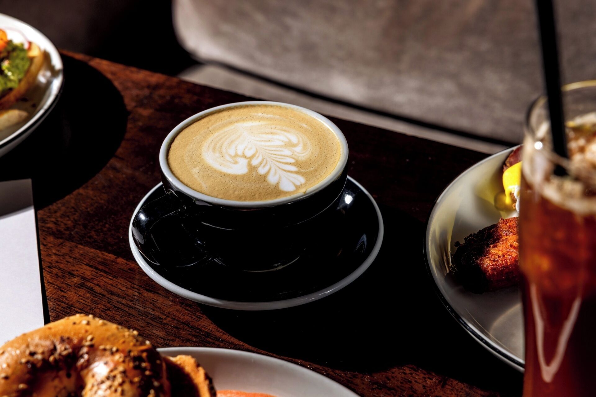 A cup of coffee with latte art on a saucer, a plate with a food item, and a glass of a beverage with a garnish.