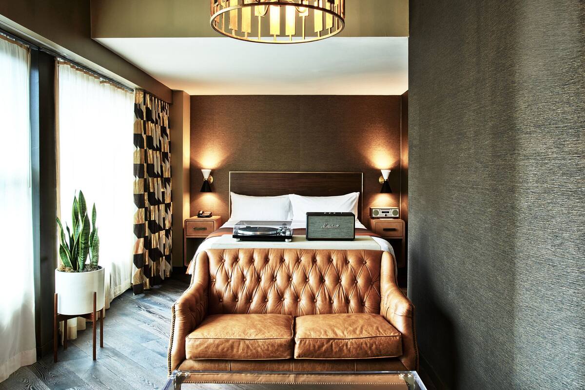 A hotel room with a tufted leather sofa, a bed with a wooden headboard, bedside lamps, a radio, and a potted plant.
