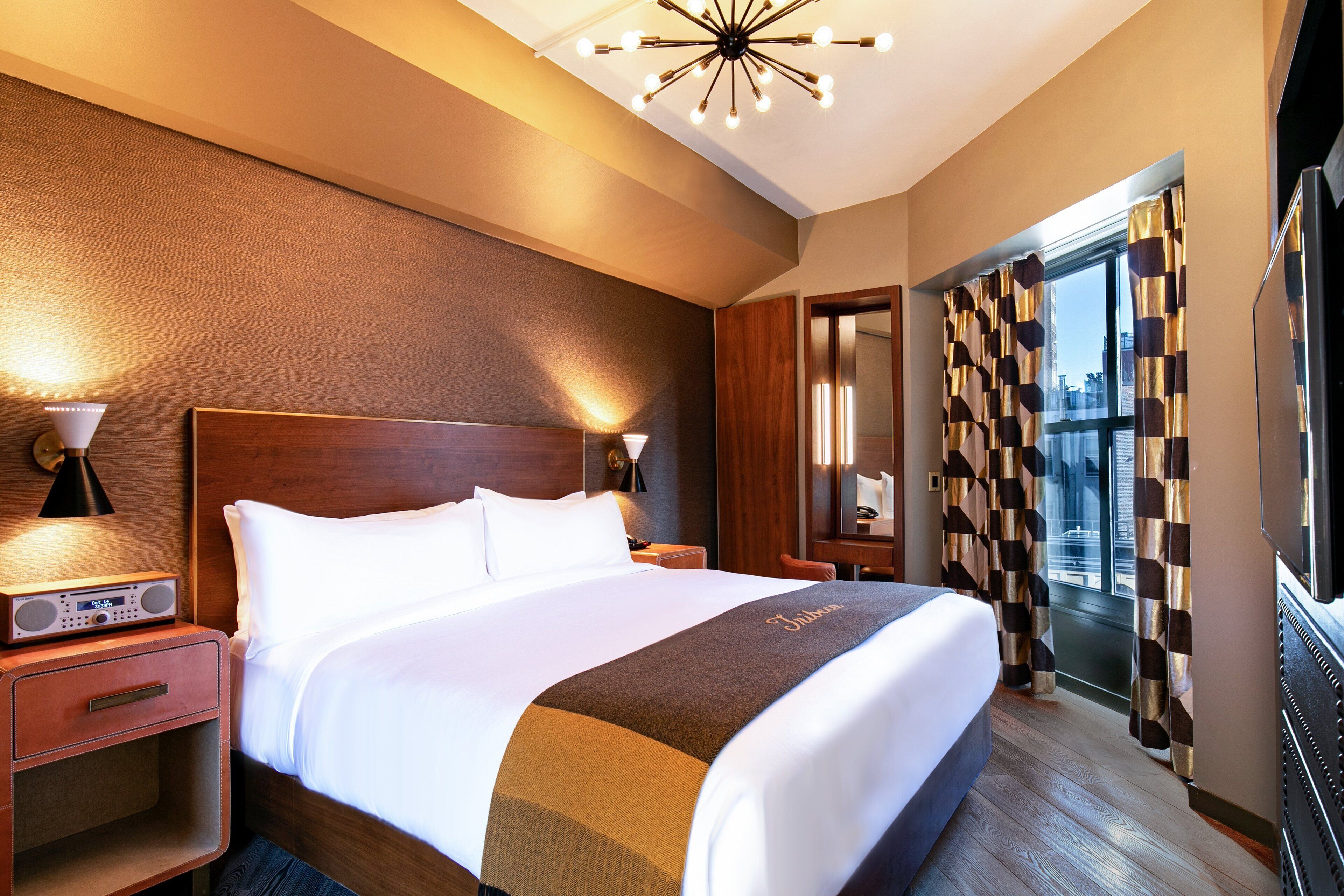 A hotel room with a large bed, wooden headboard, bedside tables with lamps, a radio, and a view of the city through large windows with patterned curtains.
