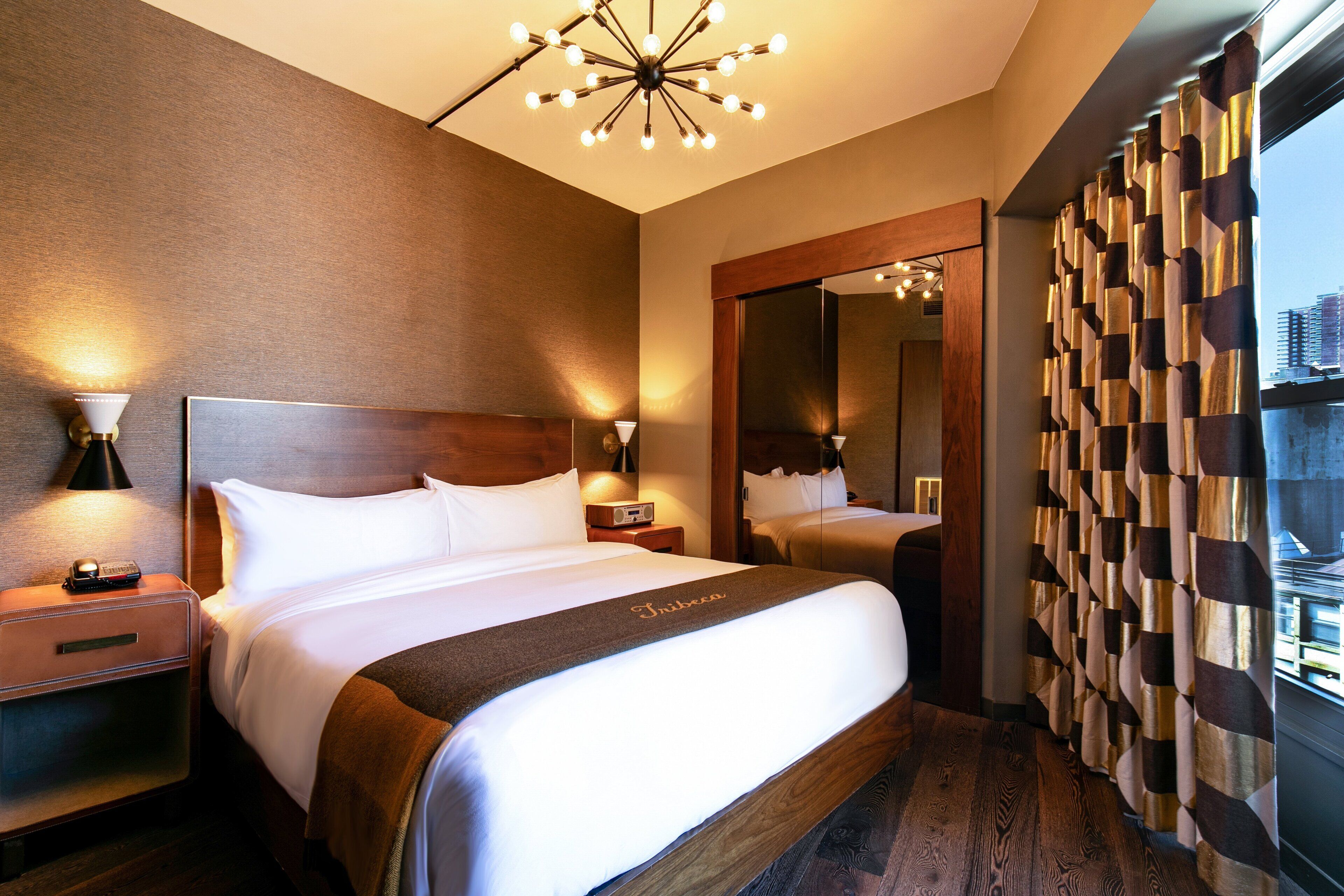 A hotel room with a large bed, a nightstand with a lamp, a mirror, and a view of the city through the window.