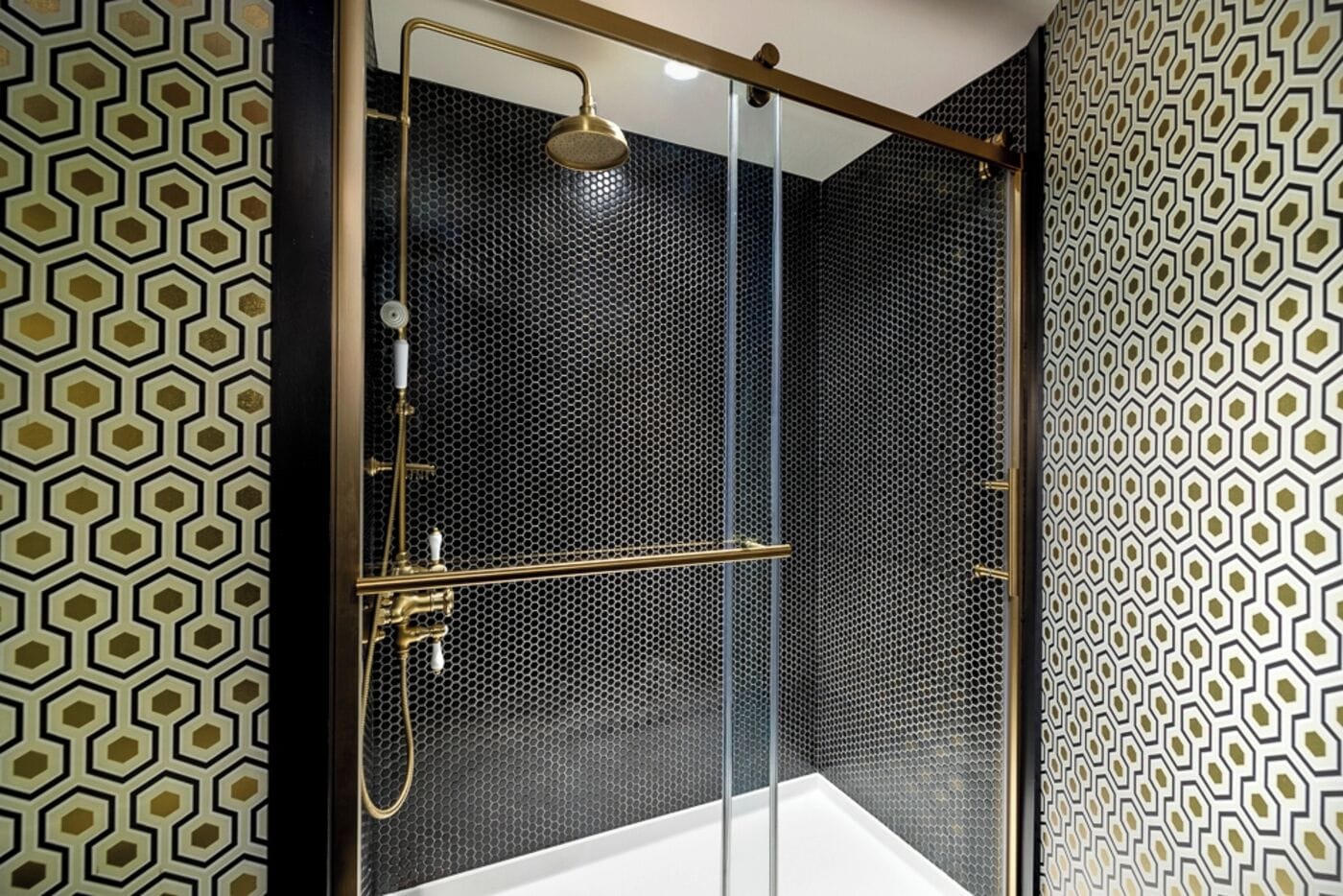 A modern bathroom with a glass-enclosed shower, black hexagonal patterned walls, and a gold showerhead.