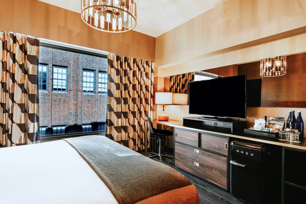 A hotel room with a bed, a desk with a TV, a minibar, and a view of a brick building.