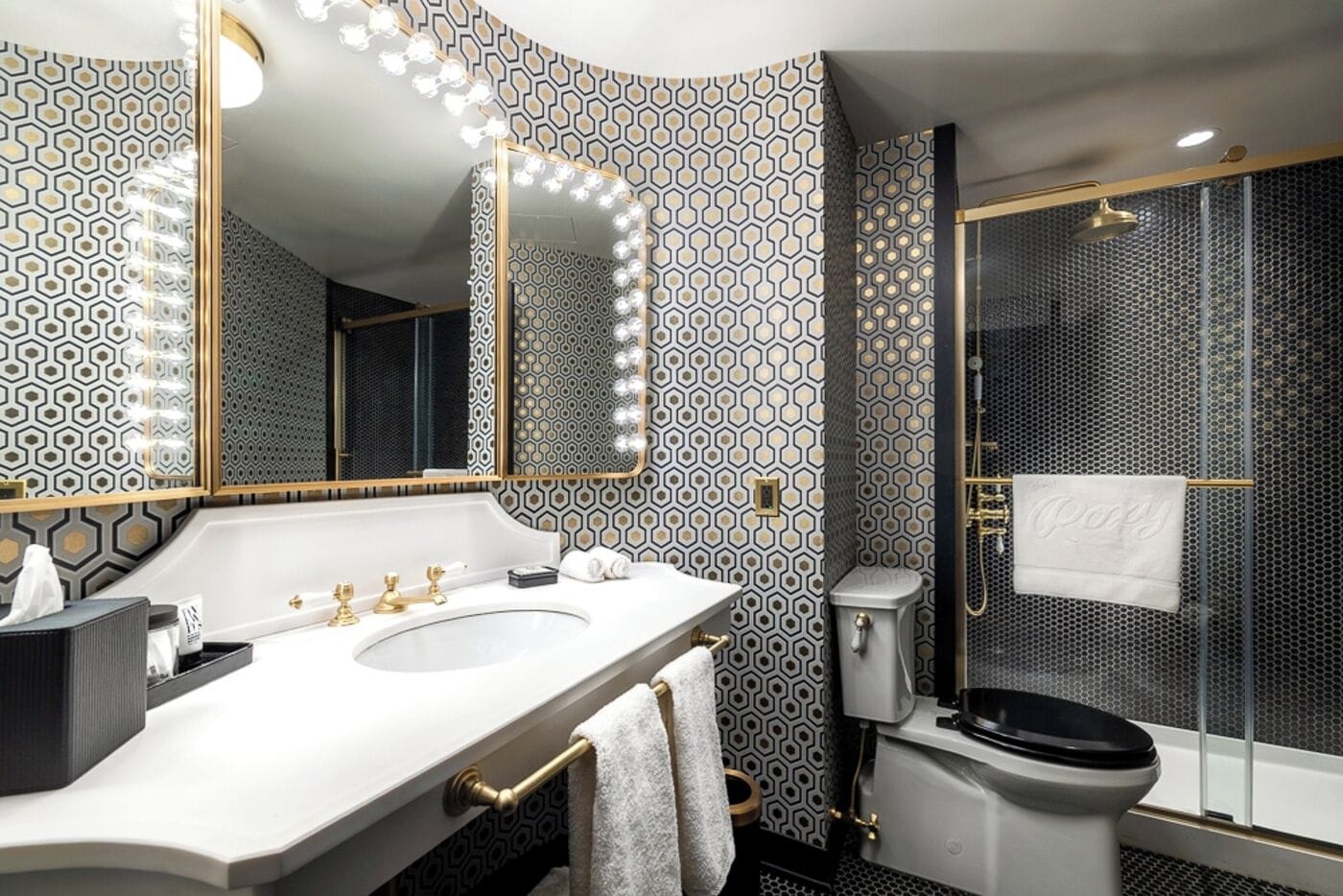 A modern bathroom with a dual sink vanity, a large mirror with lights, a shower enclosure, and patterned wallpaper.