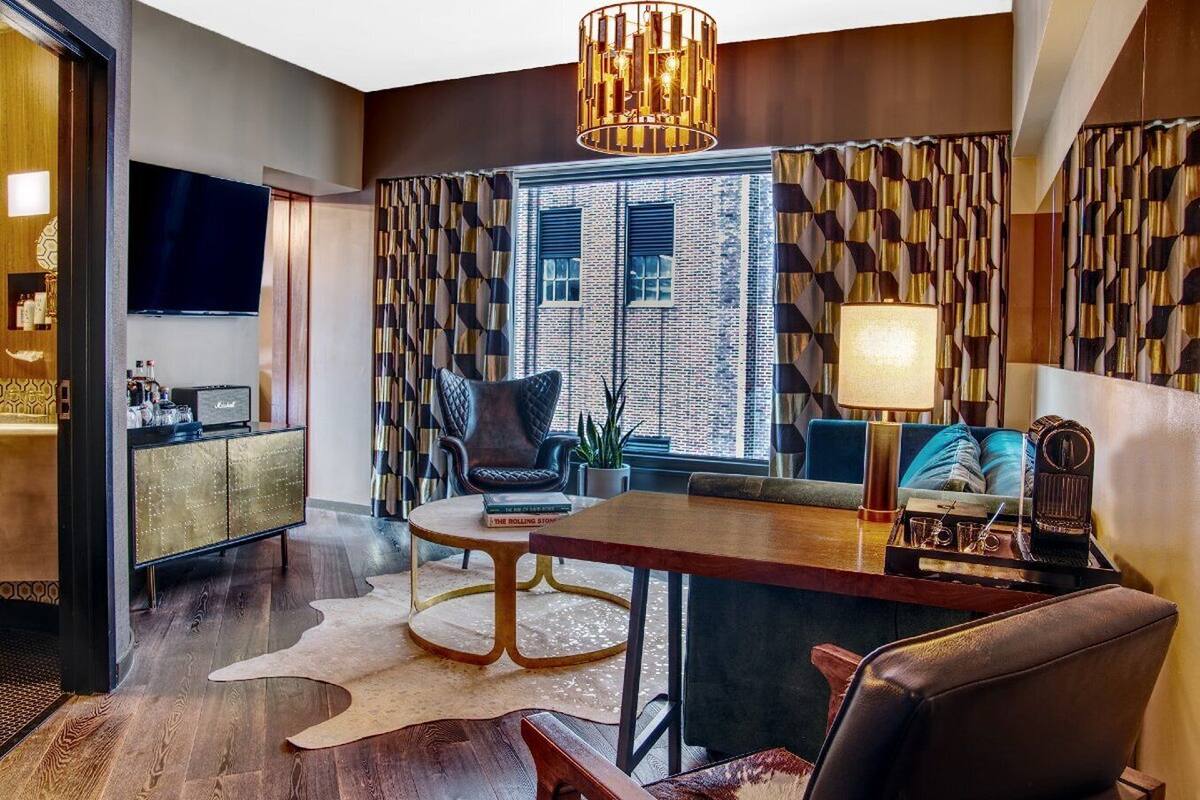 A modern living room with a large wooden dining table, a comfortable sofa, a black leather armchair, and a television on a wooden cabinet.