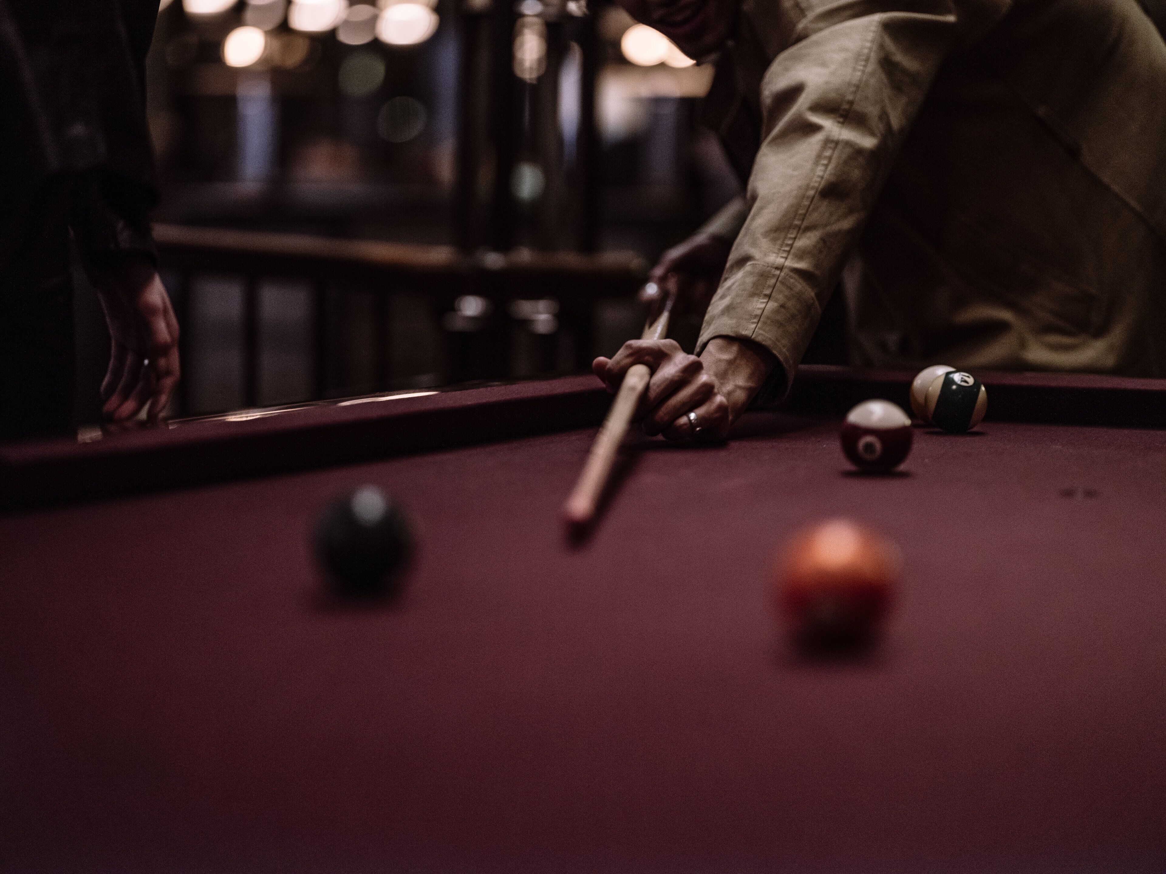 A close-up of a pool table with a cue stick, balls, and a person's hand reaching for a ball.