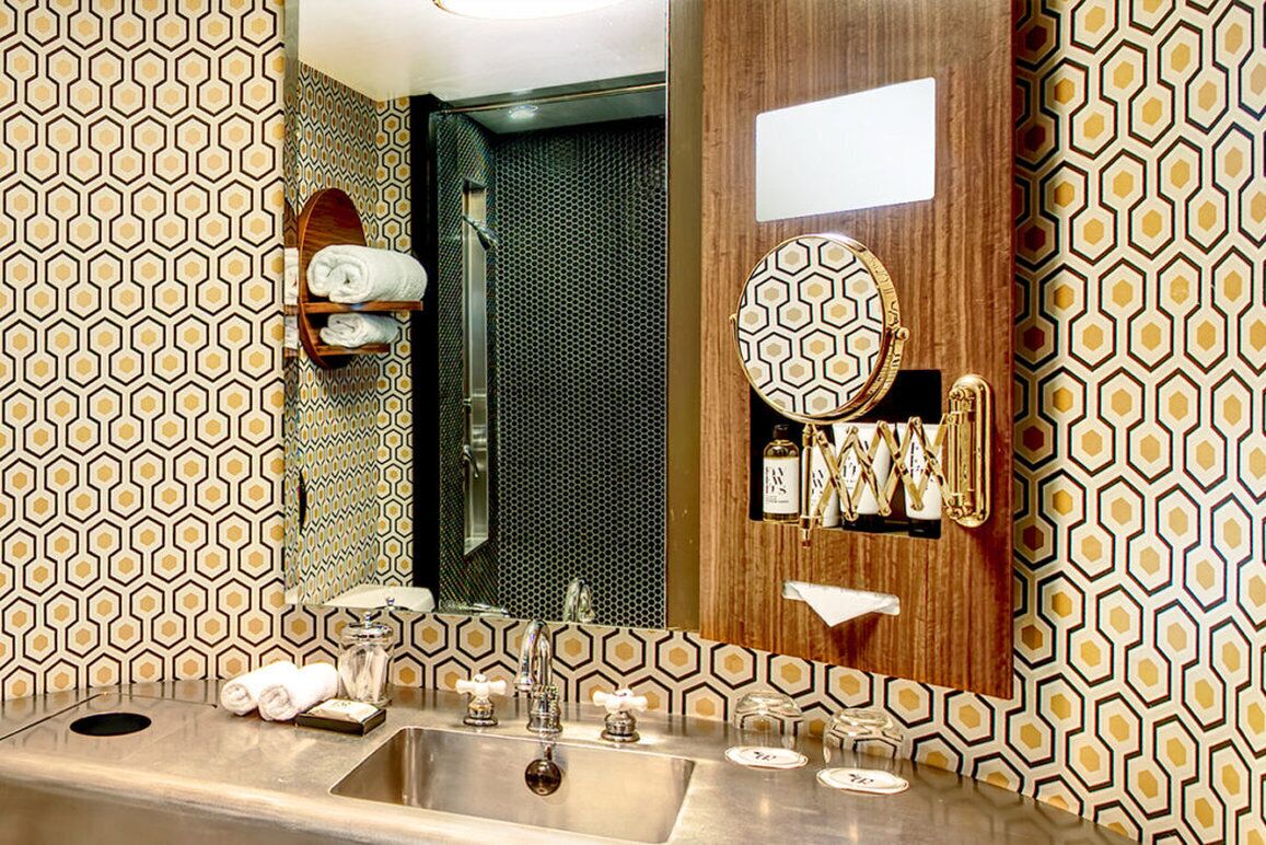 A bathroom with a geometric patterned wall, a wooden vanity, a mirror, a sink with two faucets, and a towel rack.
