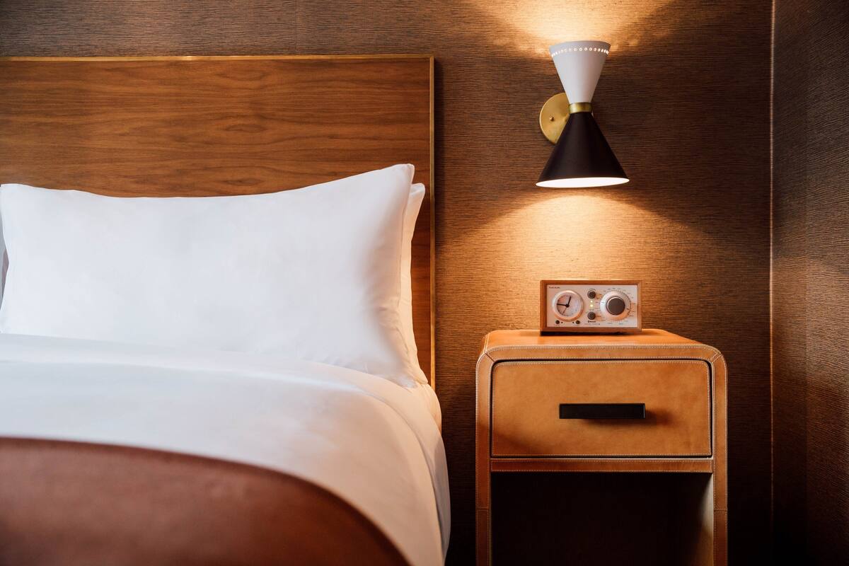 A neatly made bed with white linens, a wooden headboard, a bedside table with a radio, and a wall-mounted lamp.