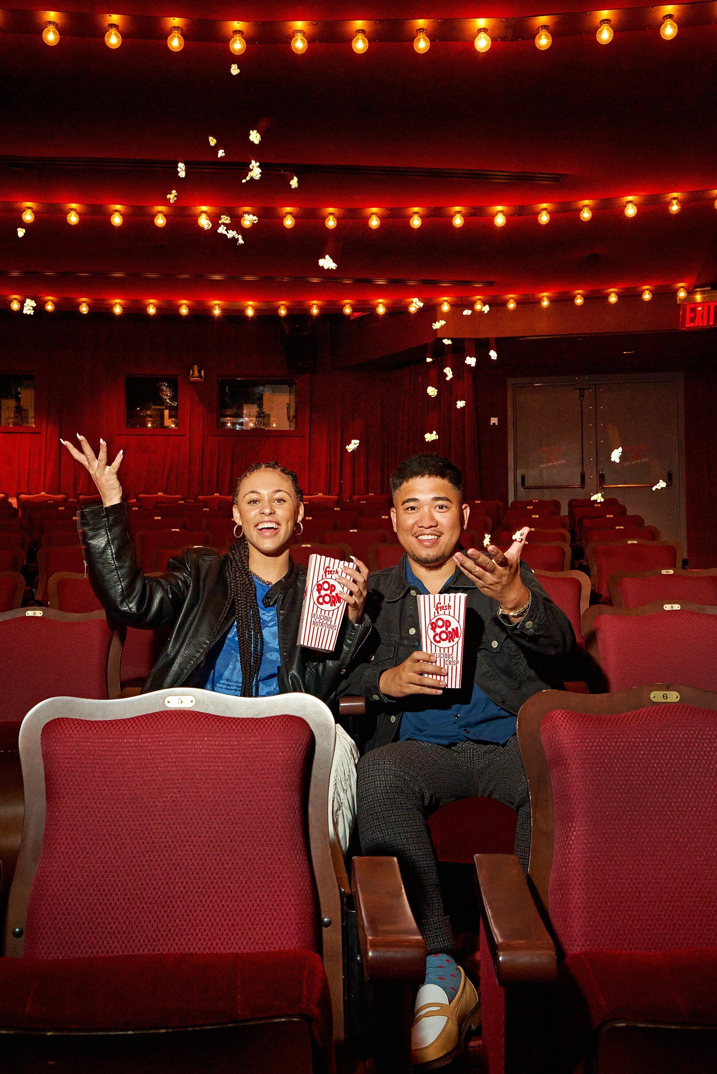 Two people seated in a theater with red seats and popcorn boxes.