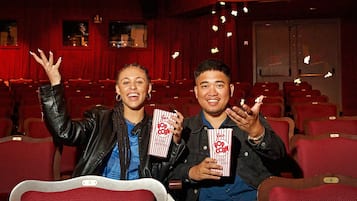 Two people seated in a theater with red seats and popcorn boxes.