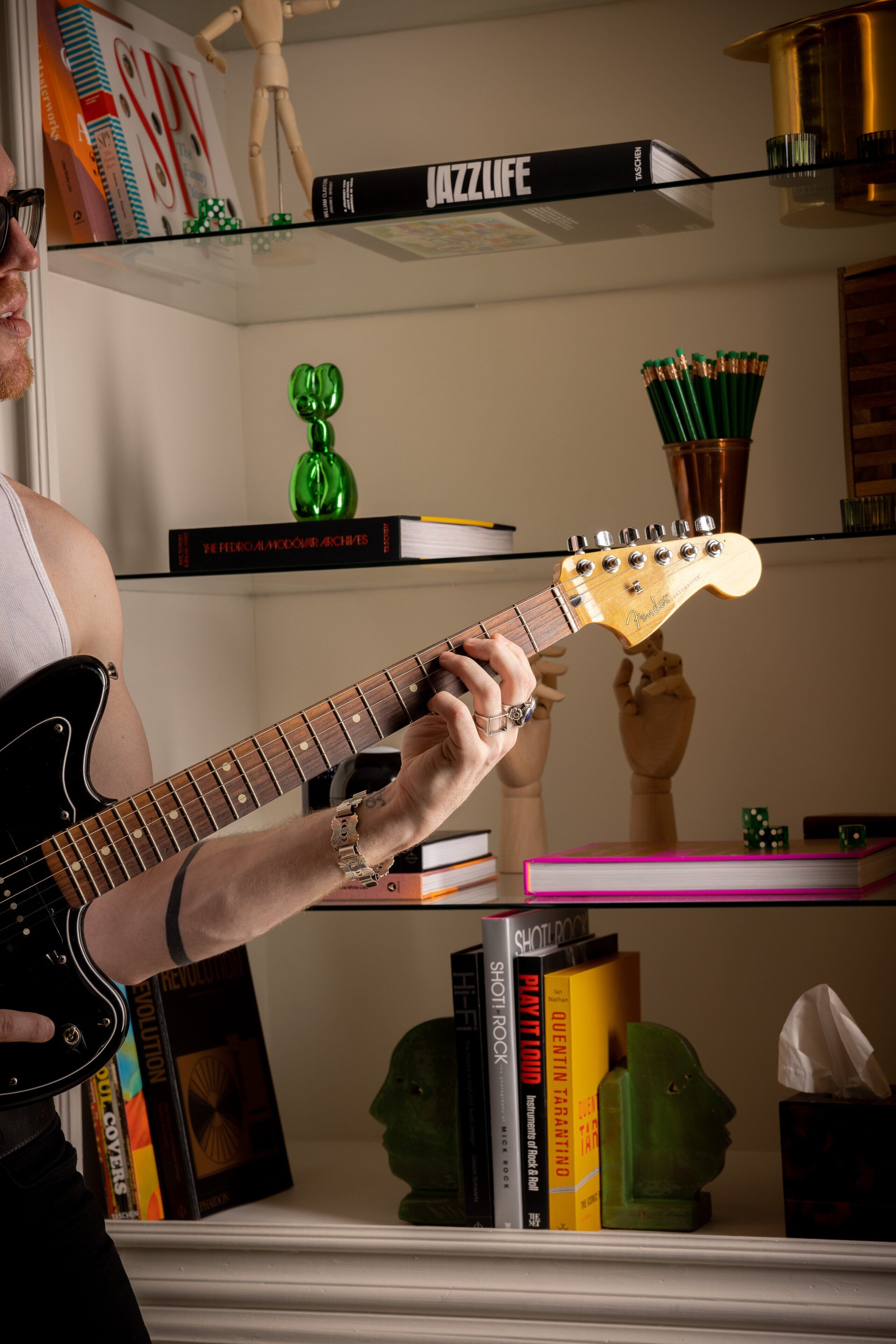 A person playing an electric guitar in a room with bookshelves, a green sculpture, and a wooden bust on display.