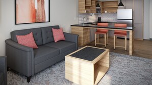 Una sala de estar moderna con un sofá gris, una mesa de centro de madera, y una zona de cocina con una lámpara colgante negra, armarios de madera y un refrigerador blanco.