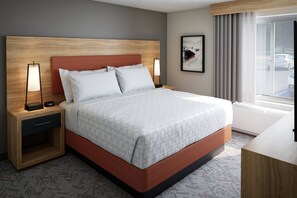 A hotel room with a bed, nightstands, a lamp, a clock, a painting, and a window with a view of cars. - Candlewood Suites Houston NE - Stafford by IHG (Houston)