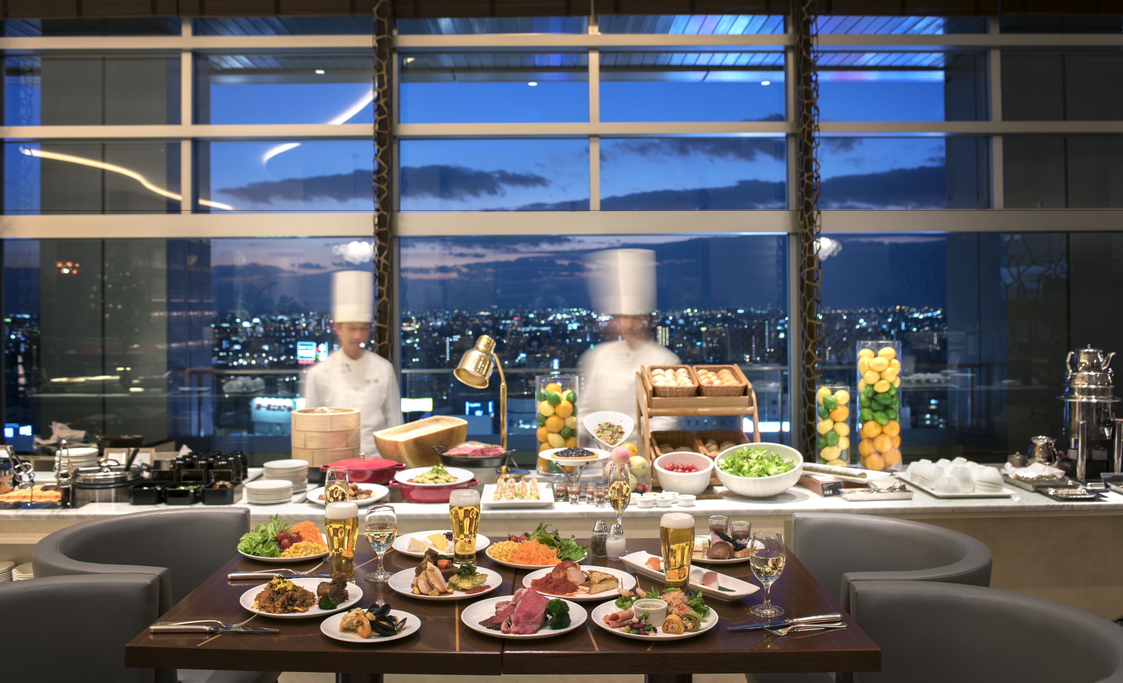 A dining table set with various dishes and drinks, a cityscape view through large windows, and two chefs in the background.