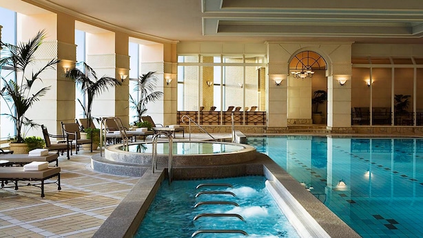 Una piscina interior con sillas de descanso y tumbonas, rodeada de grandes ventanales y plantas en macetas.