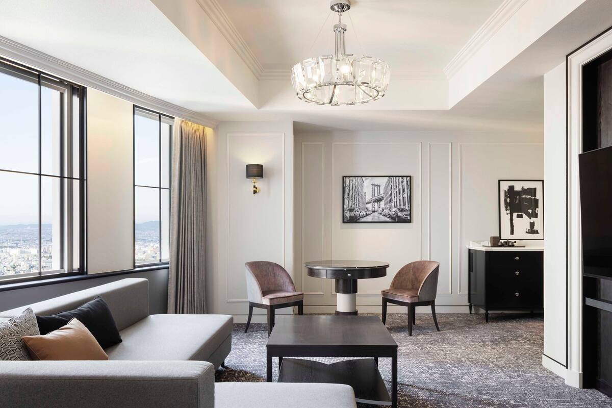 A modern hotel room with a large window, a sofa, a dining table, two chairs, a small cabinet, and a chandelier.