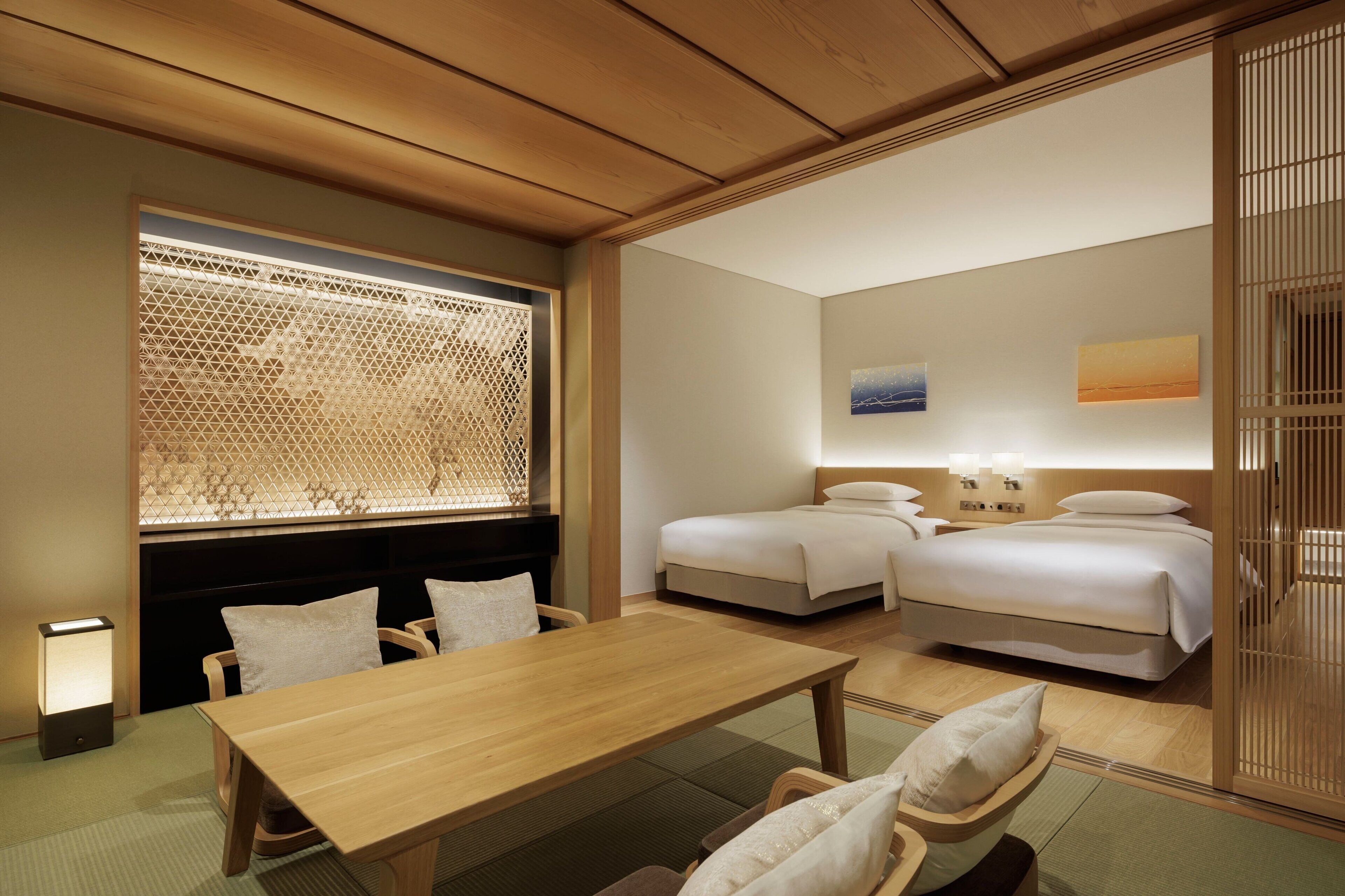 A hotel room with a wooden ceiling, a dining table, two beds, and a window with a decorative screen.