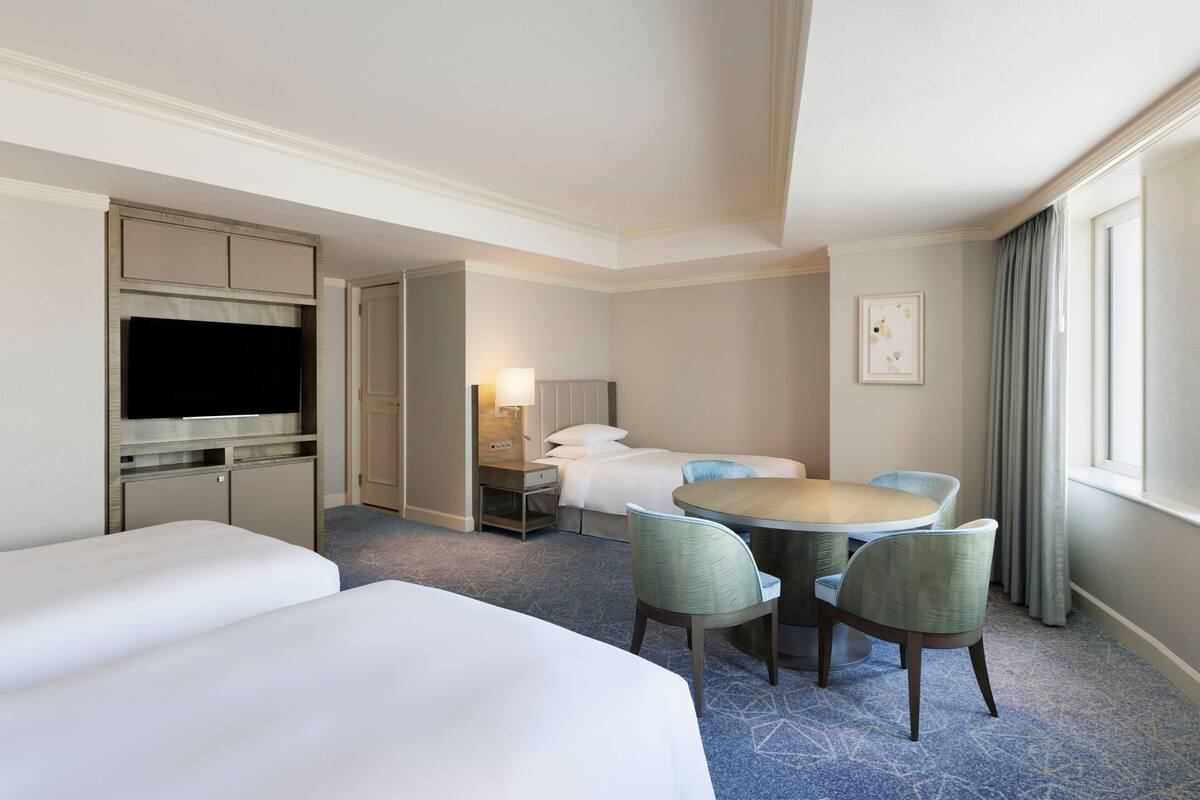 A hotel room with a bed, a TV, a desk with a lamp, and a dining area with chairs.