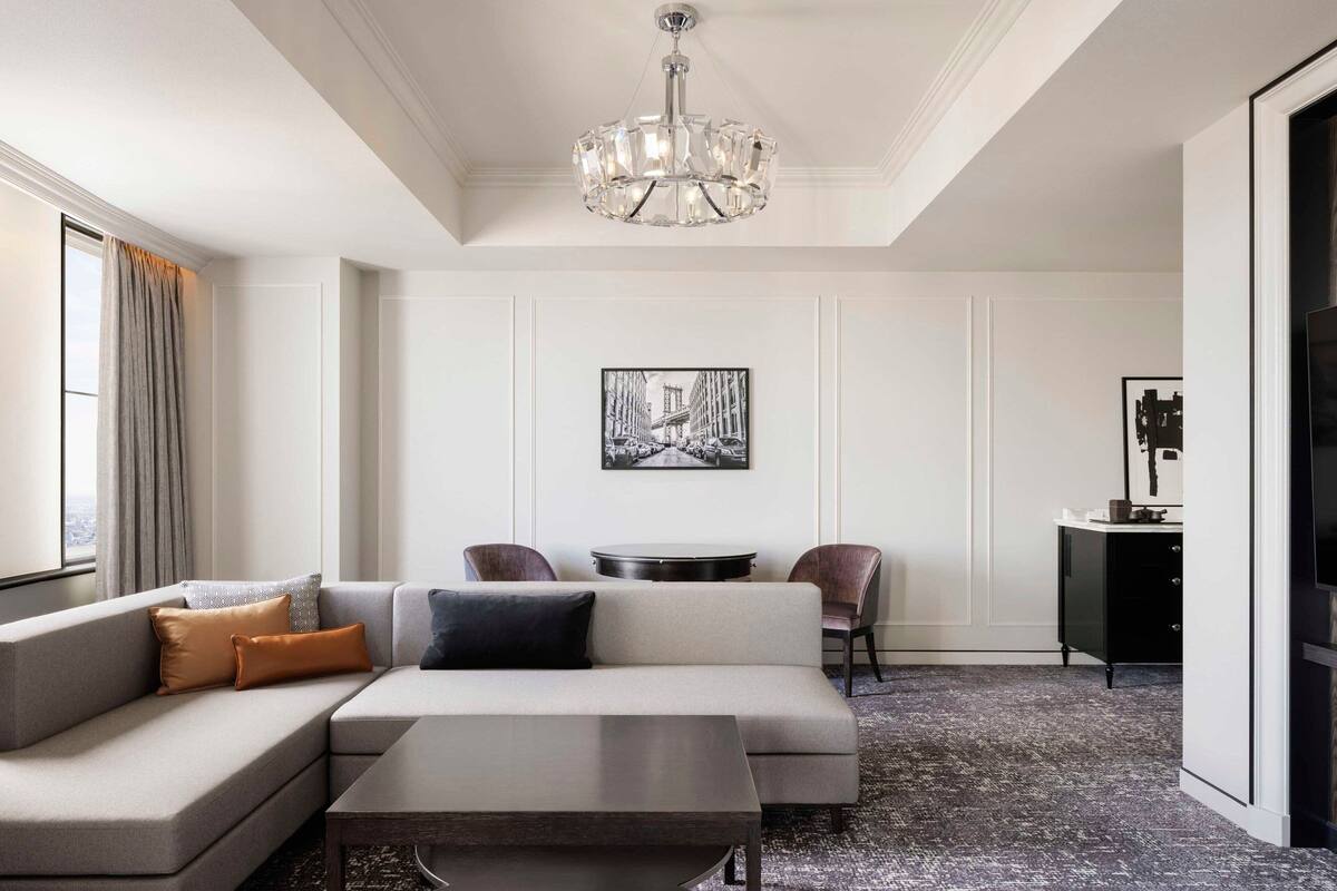 A modern living room with a sectional sofa, a coffee table, and a chandelier. There is a dining area with a round table and chairs, and a painting on the wall.