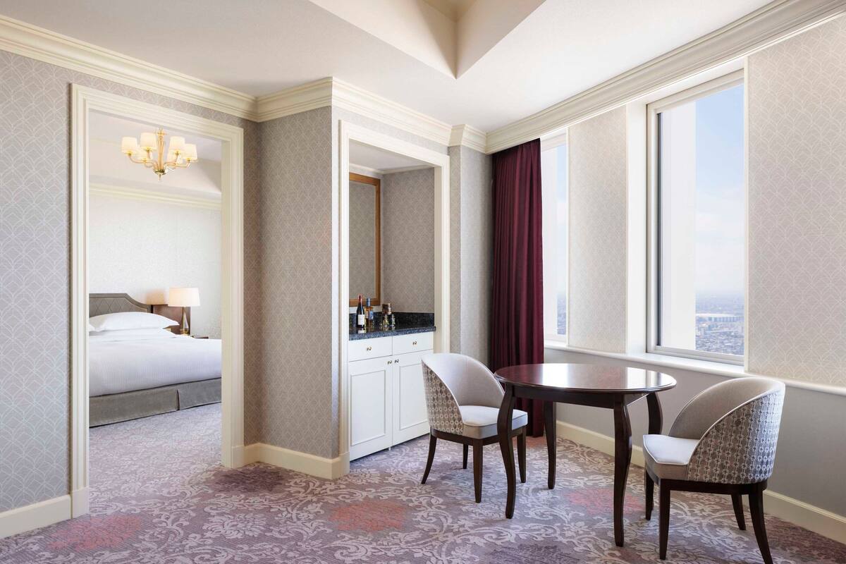 A hotel room with a bed, a desk with a chair, a large window with a view, and a patterned carpet.