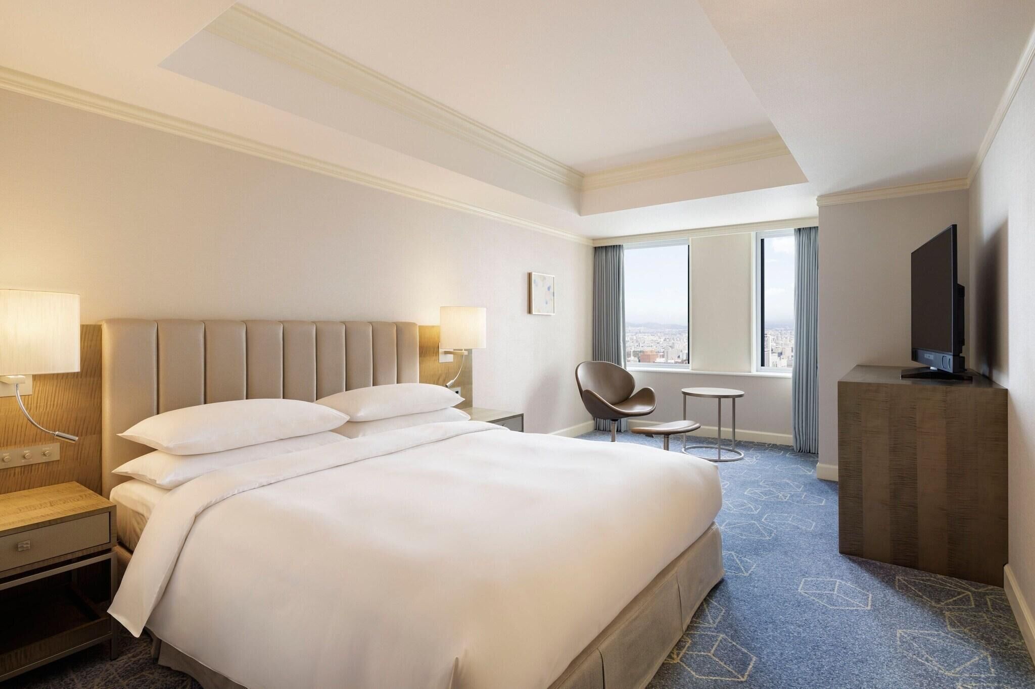 A hotel room with a large bed, a desk, a chair, a TV, and a window with a city view.