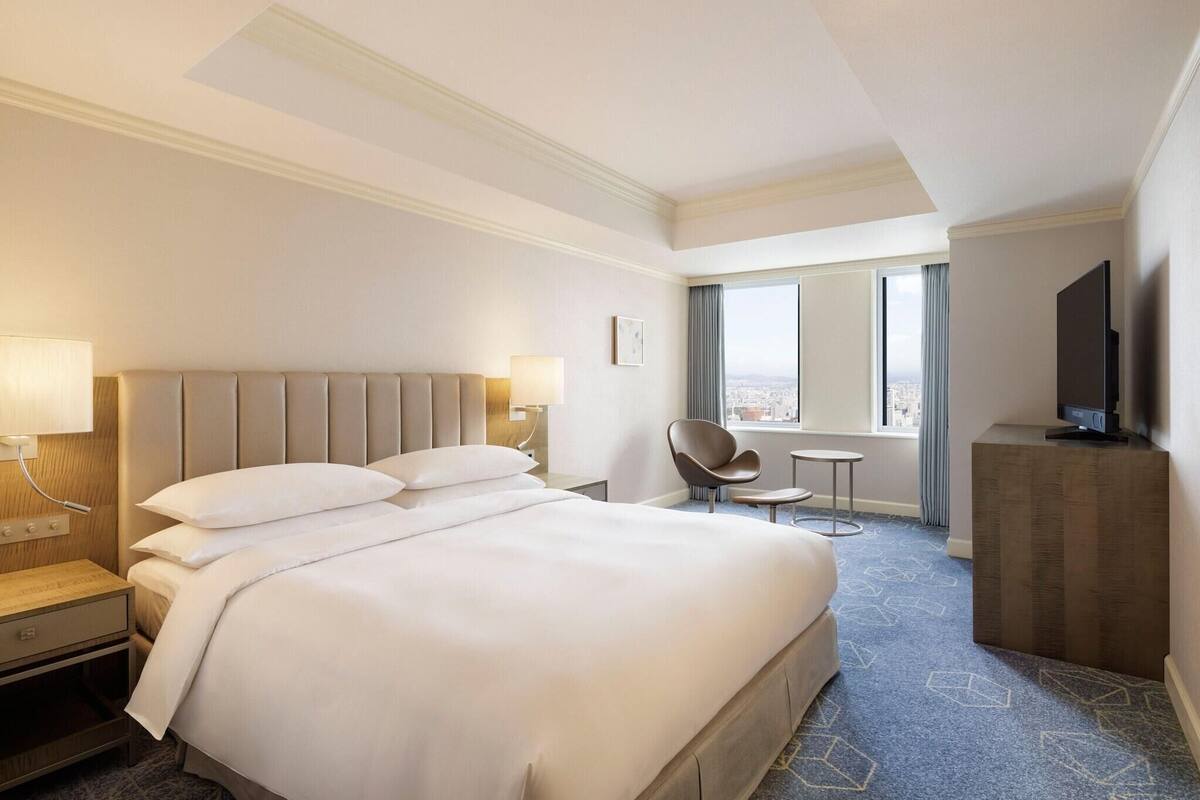 A hotel room with a large bed, a desk, a chair, a TV, and a window with a city view.